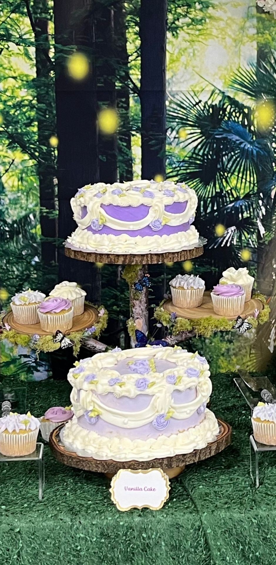 Tiered vanilla cake and cupcakes with purple and white frosting in a forest-themed setting.